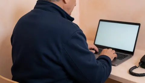 Homme adulte utilisant un ordinateur portable dans un bureau moderne aux murs en plâtre terracotta, portant une veste bleu foncé, évoquant un environnement professionnel pour un cours de programmation.