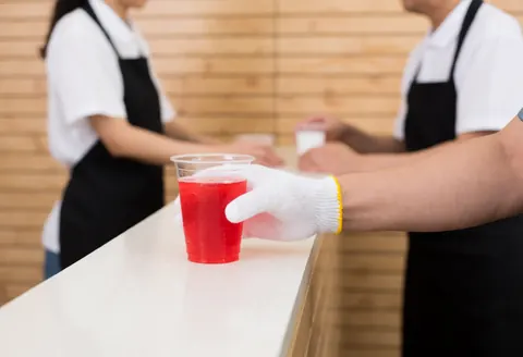 Une main portant un gant blanc dépose un verre de plastique contenant une boisson rouge sur un comptoir en bois clair dans un restaurant de restauration rapide.