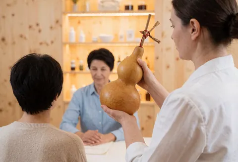 Un praticien tient un gourde avec des tiges pendant qu'il sert un client assis à une table, avec des étagères en bois et des murs en arrière-plan.