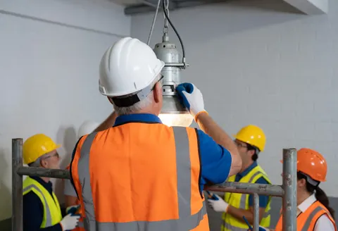 Un électricien ajuste un projecteur industriel suspendu à un échafaudage métallique dans un entrepôt, avec d'autres travailleurs en arrière-plan.