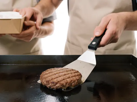 Cuisinier portant un tablier servant un hamburger sur une plaque en acier avec une spatule, fumée visible dans un restaurant de restauration rapide.