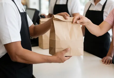 Employé de restaurant remet un sac de repas à un client derrière un comptoir moderne et propre dans un environnement professionnel.