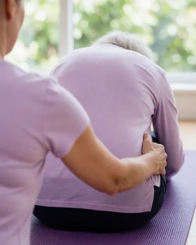 Instructrice aidant une cliente âgée en posture de flexion sur une manteau violet dans un studio lumineux avec fenêtre.