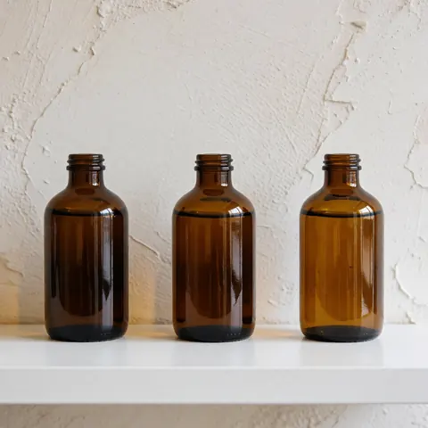 Trois bouteilles en verre marron contenant de l'huile de massage cervical posées sur une étagère blanche devant un mur texturé avec une lumière naturelle.