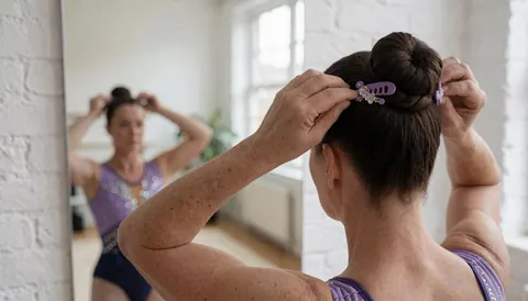Gymnaste professionnelle ajustant sa coiffure avec une épingle décorative devant un miroir, sur fond de mur de briques blanches, portant un justaucorps violet pailleté.
