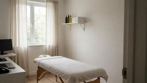Salle de massage avec table recouverte d'un drap blanc, étagère avec serviettes et huiles, fenêtre avec rideaux et murs blancs.