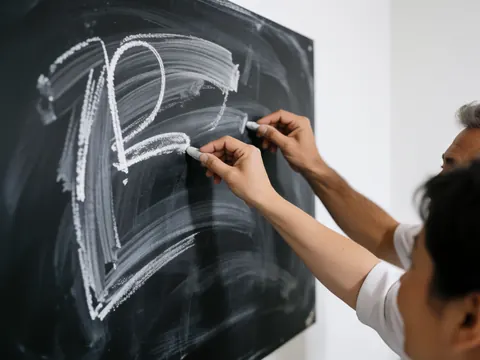 Gros plan d'une main adulte tenant une craie blanche et écrivant sur une surface de tableau noir, illustrant l'apprentissage et la formation en ligne.