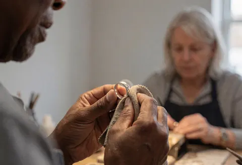 Un technicien poli une bague en argent avec un chiffon doux dans un atelier de joaillerie aux murs gris et à la lumière naturelle.