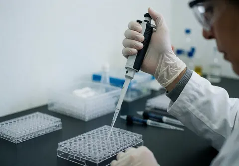 Technicien de laboratoire utilisant une micropipette pour transférer du liquide dans une microplaque transparente posée sur un plan de travail sombre.
