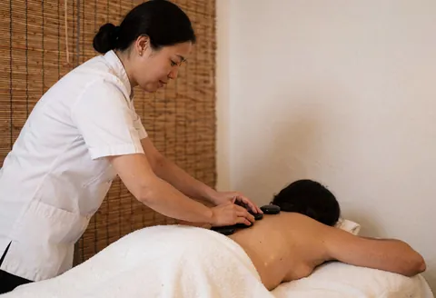 Un masseur en uniforme blanc pose des pierres chaudes sur le dos d'un client allongé sur une serviette blanche dans un spa aux murs blancs et aux stores en bambou