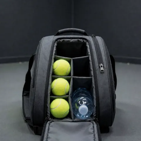 Valise de tennis ouverte contenant trois balles jaunes et une bouteille d'eau, posée sur une surface grise, évoquant la préparation d'un instructeur de tennis.