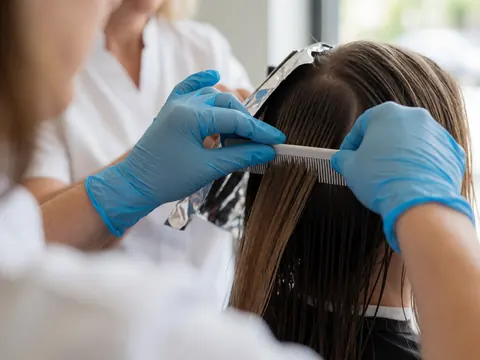 Des mains portant des gants bleus peignent des cheveux humides sur une feuille métallique dans un salon de coiffure.