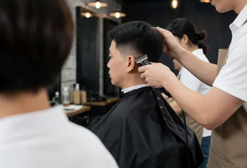 Un barbier professionnel utilise une tondeuse pour couper les cheveux d'un client dans un salon aux murs sombres et aux lumières industrielles.