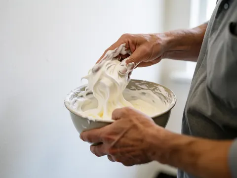Mains d'un boulanger mélangeant une meringue crémeuse dans un bol métallique sur un fond blanc avec une lumière vive.