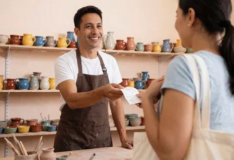 Un vendeur souriant remet un reçu à un client tenant une pochette en toile, devant des étagères remplies de céramiques colorées dans un atelier artisanal.