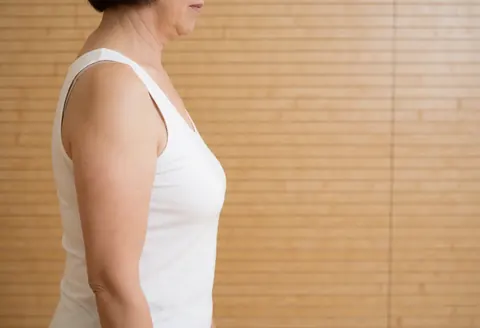 Femme adulte en profil latéral, portant un t-shirt blanc, devant un mur de planches de bois clair, éclairée par la lumière naturelle.