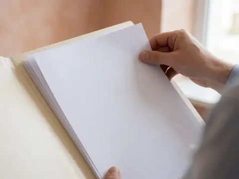 Une main range des papiers blancs dans un classeur sur un bureau, avec un mur terracotta en arrière-plan et une lumière naturelle douce.