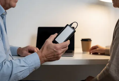 Une main tient un disque dur externe relié par câble, dans un bureau avec un ordinateur portable et une tasse de café, évoquant un transfert de données.