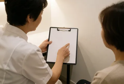 Un professionnel en blouse blanche explique des soins à un client assis dans un cabinet lumineux aux murs blancs.