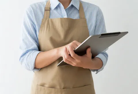 Personne portant un tablier et une chemise à rayures tenant une tablette numérique sur fond blanc, évoquant les services professionnels et le soutien technique.