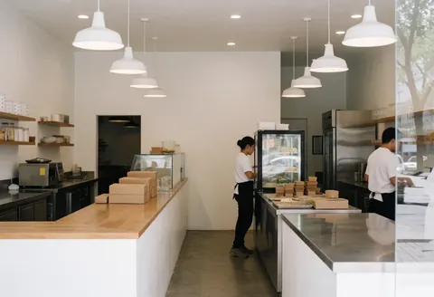 Deux employés servent des gâteaux dans une pâtisserie moderne avec des comptoirs en acier inoxydable et des suspensions blanches.