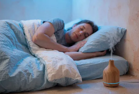Une personne dort paisiblement dans un lit près d'un diffuseur émettant de la vapeur, sur fond de murs terracotta et d'une lumière bleue douce.