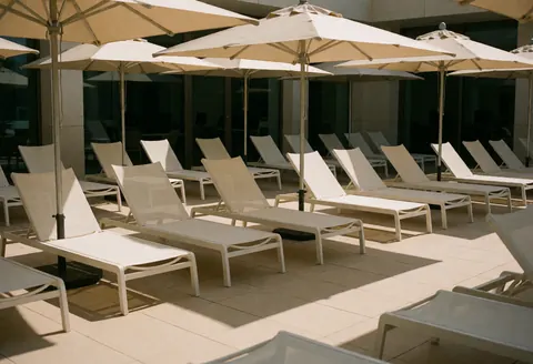 Scène de piscine publique avec chaises de plage blanches et parasols beige sous un soleil éclatant, idéale pour se détendre en plein air.
