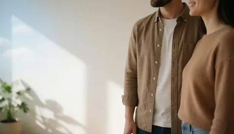 Un couple sourit près d'un mur blanc éclairé par la lumière du soleil, avec l'ombre d'une plante, créant une ambiance calme et naturelle.