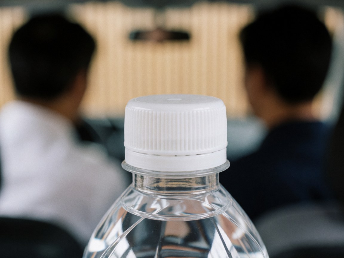 Gros plan sur le bouchon d'une bouteille d'eau avec une texture visible, sur un fond flou montrant deux personnes et des murs en bois clair, idéal pour illustrer la propreté ou l'hygiène.