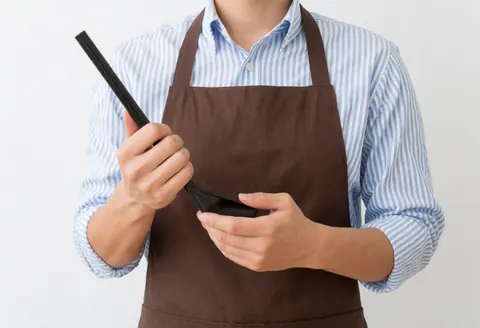 Personne vêtue d'un tablier tenant une cuillère de cuisine sur un fond blanc, évoquant un environnement de travail culinaire.