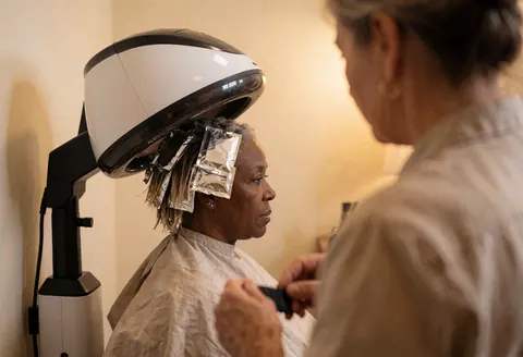 Une cliente portant des mèches de coloration sous un sèche-cheveux dans un salon de coiffure aux murs beiges et à la lumière chaleureuse.