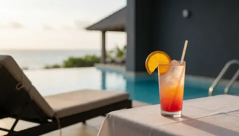 Cocktail avec tranche d'orange sur table blanche près d'une transat et d'une piscine avec vue sur la mer, idéal pour un resort touristique.