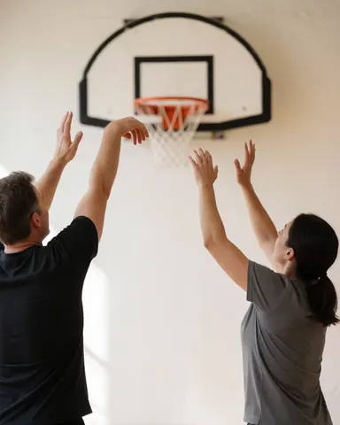 Deux individus pratiquant le basketball dans un gymnase aux murs blancs et à la lumière naturelle, avec un panier visible.