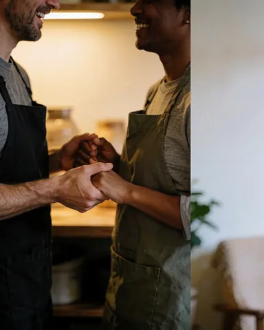 Deux personnes portant des tabliers sourient et se tiennent par la main dans une cuisine éclairée de lumière chaude, illustrant le travail d'équipe.
