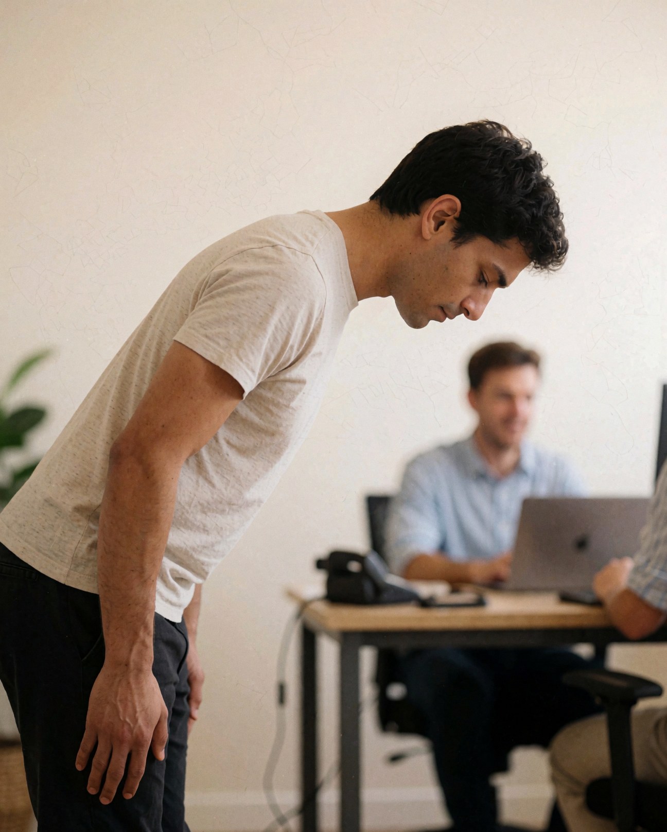 Un homme debout salue dans un bureau moderne avec un collègue travaillant sur un ordinateur portable en arrière-plan.
