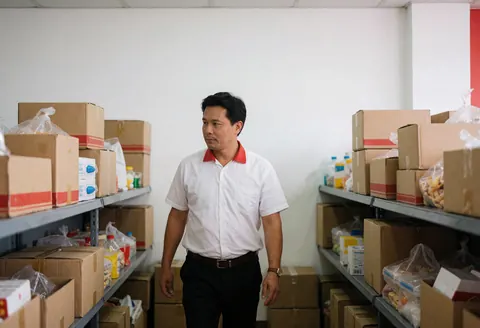 Un manager se déplace parmi des étagères remplies de boîtes et de produits emballés dans un entrepôt de restauration rapide aux murs blancs et à l'éclairage fonctionnel.