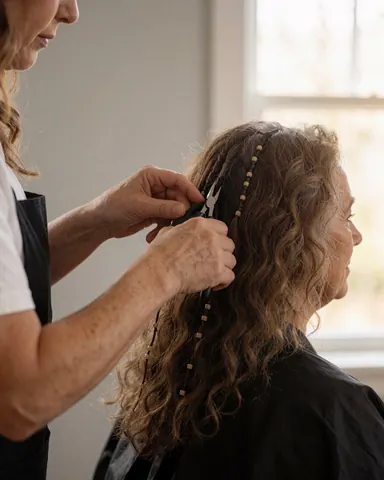 Une coiffeuse utilise des pinces pour fixer des extensions sur les cheveux bouclés d'une cliente assise près d'une fenêtre baignée de lumière naturelle.