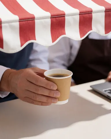 Une main tenant une tasse de café posée sur une table en extérieur, à l'ombre d'un store rouge et blanc, évoquant la détente.