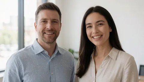 Deux personnes souriantes regardant l'objectif, vêtues de chemises à manches longues, dans un bureau moderne baigné de lumière naturelle.