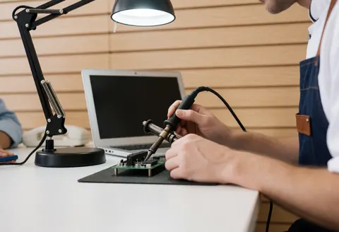 Technicien en train de souder une carte électronique à l'aide d'une lampe de bureau dans un atelier de robotique avec un mur en bois clair et du matériel moderne.