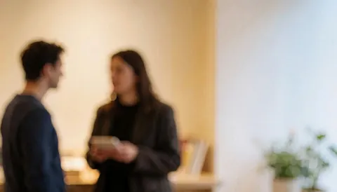 Deux individus en conversation dans un espace intérieur aux murs clairs et à l'éclairage doux, offrant un fond neutre adapté à l'ajout de texte.