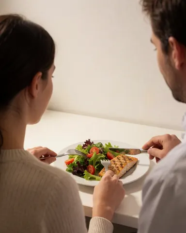 Une cliente savoure du poisson grillé et une salade sur une table blanche, avec un entraîneur de fitness en arrière-plan, dans une ambiance saine et professionnelle.