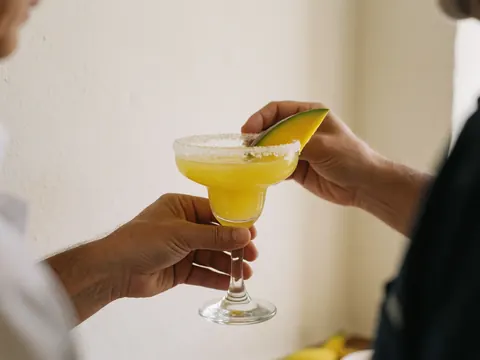 Mains tenant un verre de margarita bordé de sel et garni de mangue sur fond de mur blanc chaud, boisson rafraîchissante pour club de plage.