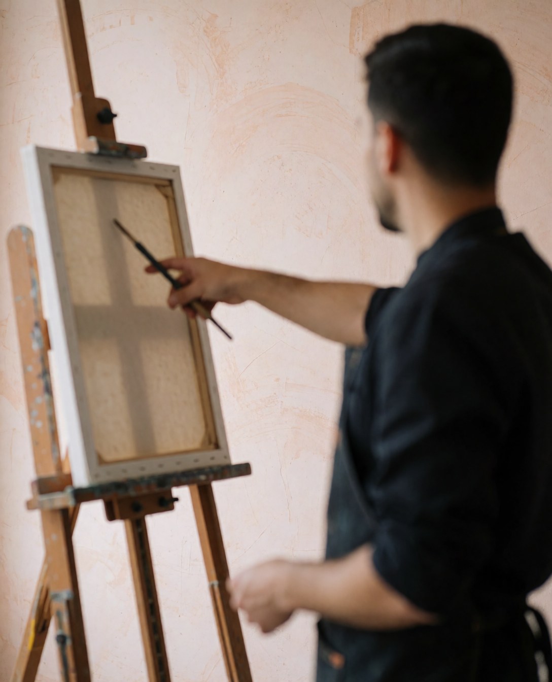 Vue de dos d'un artiste travaillant sur une toile posée sur un chevalet en bois dans un atelier aux murs clairs en plâtre.