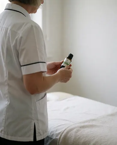 Thérapeute en salle de massage avec lit blanc et huile essentielle à la main, préparant un traitement.
