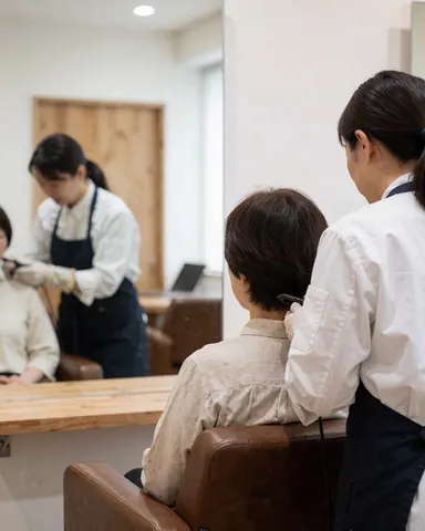 Une coiffeuse coupe les cheveux d'une cliente assise sur une chaise de salon, reflet dans un miroir, murs blancs et porte en bois.