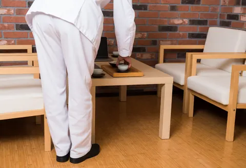 Instructeur de yoga en uniforme blanc servant du thé sur une table en bois dans une salle de repos avec mur en briques et chaises confortables.
