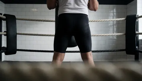 Vue de dos d'une personne dans un ring de boxe avec des murs carrelés blancs, idéale pour un site web d'académie de sport.