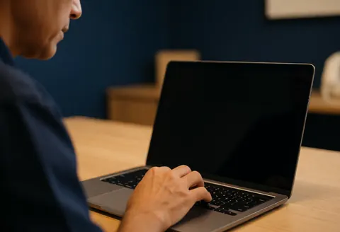 Plan rapproché d'une main posée sur le clavier d'un ordinateur portable dans un studio, avec un mur bleu foncé en arrière-plan et une lumière chaude.