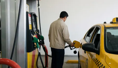 Un conducteur effectue des réparations sur un taxi jaune dans une station-service, devant un mur blanc, sans visage visible.